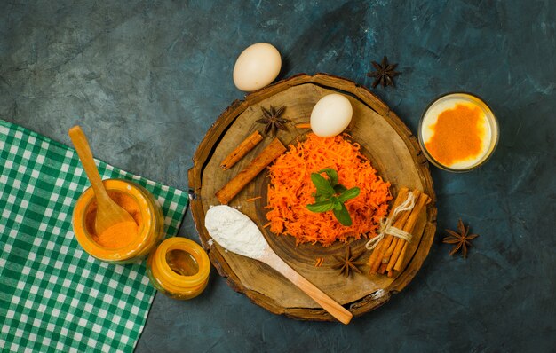 Тертая морковь с яйцами, мукой, специями, молоком, тканью для пикника на деревянной доске и лепном фоне, вид сверху.