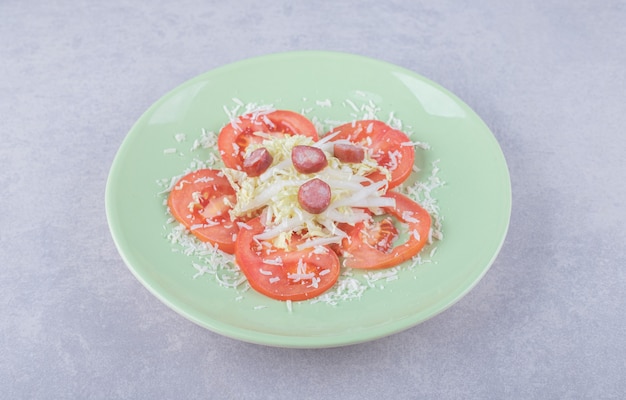 Тертый сыр с сосисками и помидорами на зеленой тарелке.