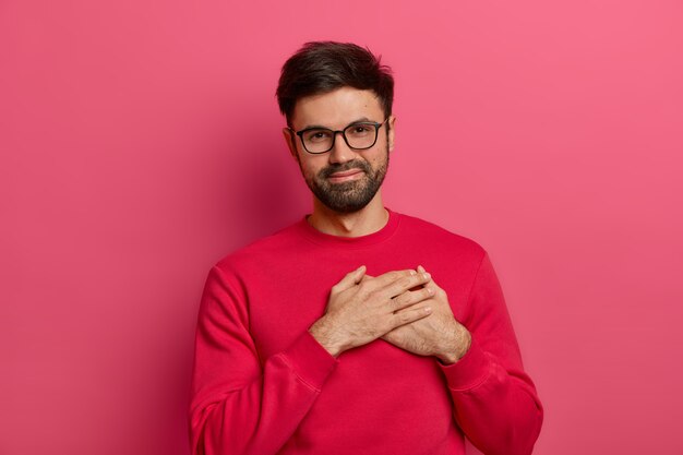 Grateful bearded man presses palms to heart, being moved and touched by pleasant words, appreciates received gift, wears spectacles and pink jumper, expresses gratitude, poses against pink wall