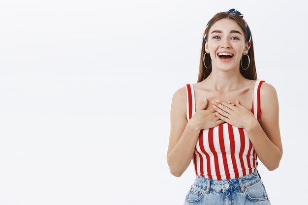 Grateful delighted young attractive woman in stylish striped top and headband holding palms on chest in thankful and pleased pose smiling broadly