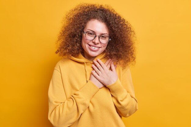 感謝の気持ちで喜んでいる巻き毛の若い女性は、心の笑顔の近くで手を握って、黄色の壁に隔離されたスウェットシャツを着て助けてくれてありがとうと言います。ボディーランゲージの概念