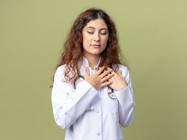 Grateful young female doctor wearing medical robe and stethoscope keeping hands on chest doing thank god gesture with closed eyes isolated on olive green wall with copy space