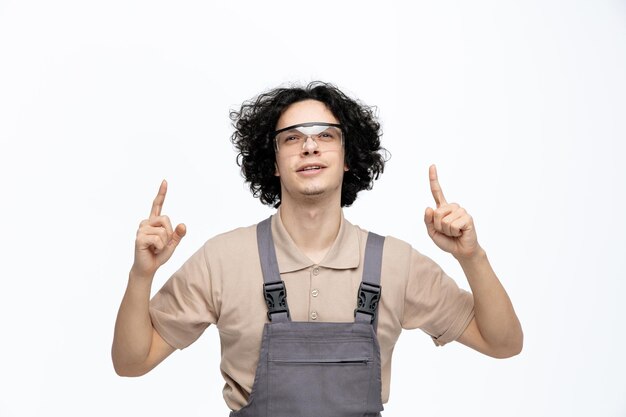 Grateful young male construction worker wearing uniform and safety glasses looking up pointing fingers up isolated on white background