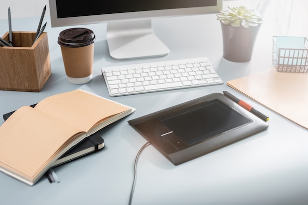 The gray desk with laptop, notepad with blank sheet, pot of flower, stylus and tablet for retouching