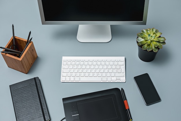 Серый письменный стол с ноутбуком, блокнотом с чистым листом, горшком с цветком, стилусом и планшетом для ретуши
