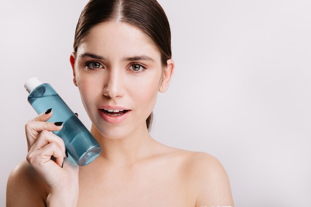 gray-eyed attractive girl with friendly smile poses without makeup and filters on white wall. lady holding blue face tonic.