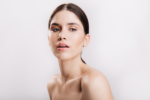 Gray-eyed female model with dark hair with healthy skin sensually posing on white wall.