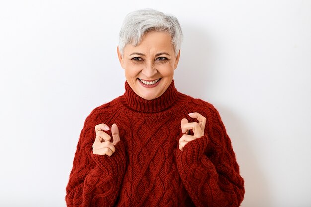 Gray haired mature senior woman in stylish knitted jumper expressing excitement and joy, looking with broad beaming smile, holding hands as if squeezing something. Human reactions and feelings