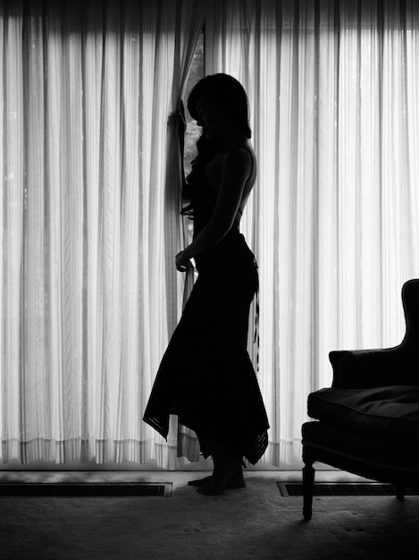 Grayscale shot of a female with an elegant dress standing near the curtain