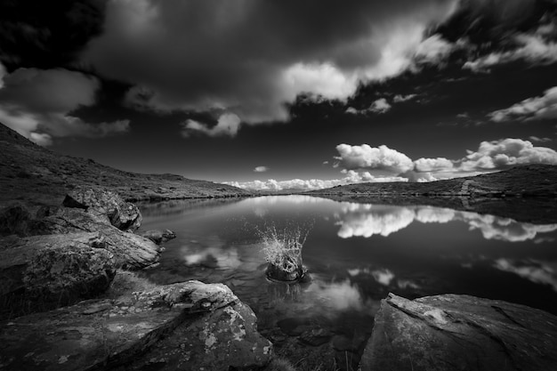 Grayscale shot of a lake surrounded by mountains under the sky full of clouds