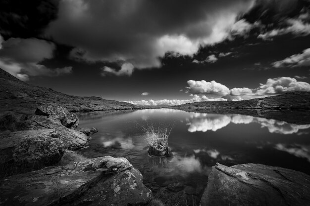 Grayscale shot of a lake surrounded by mountains under the sky full of clouds