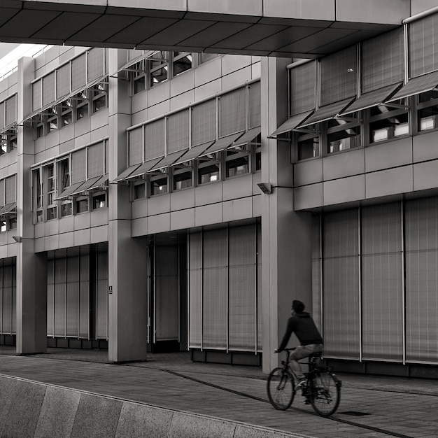 Free photo grayscale shot of a male riding a bicycle on the sidewalk next to a building with many windows