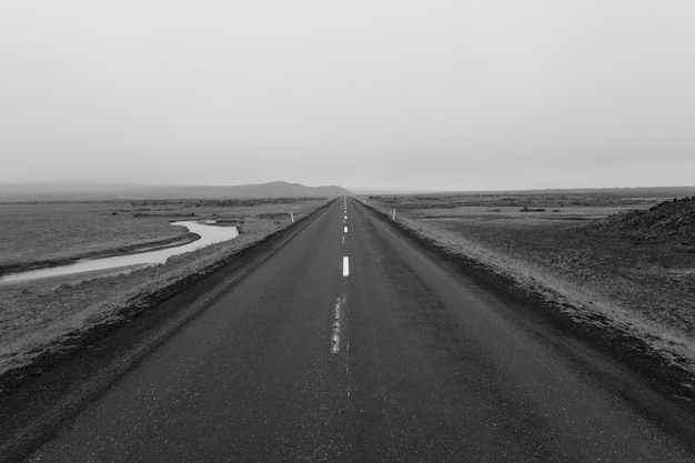 Grayscale shot of a road in the middle of an empty field under a cloudy sky