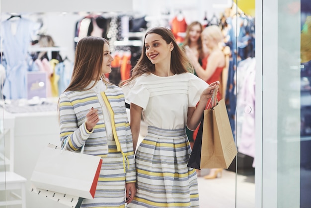 Отличный день для покупок. Две красивые женщины с сумками смотрят друг на друга с улыбкой во время прогулки по магазину одежды