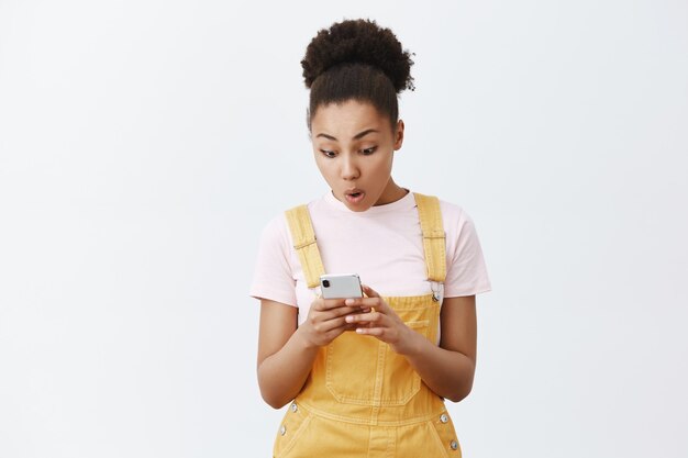 Great news from my friend. Pleased surprised and excited good-looking African American in trendy yellow overalls, holding smartphone, taping message on screen, gazing at device with amazement