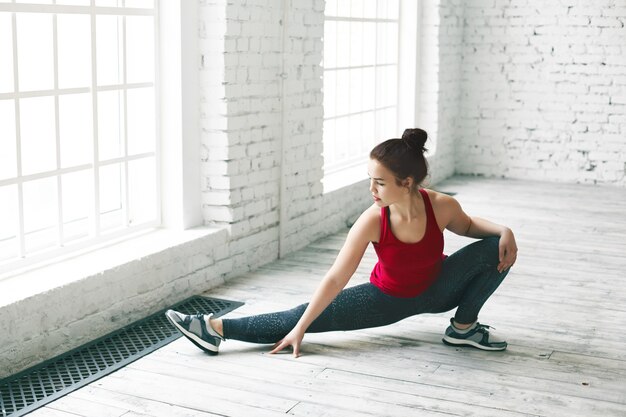 Great progress. Picture of attractive young European woman doing stretches indoors, warming up leg muscles before side splits in light hall with copy space wall for your advertising content