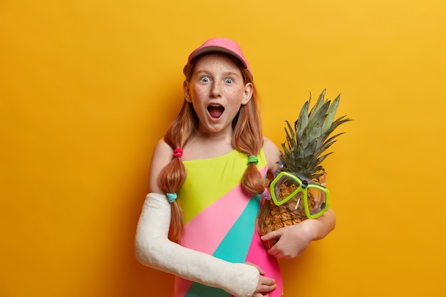 Greatly impressed freckled girl stands with widely opened mouth, embraces pineapple with snorkeling mask, enjoys summer time, has broken arm, isolated on yellow wall. Children, emotions