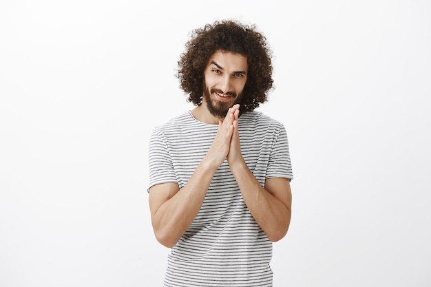 Greedy guy has nasty plan. Portrait of intrigued curly-haired man with beard, rubbing palms together near chest and smiling