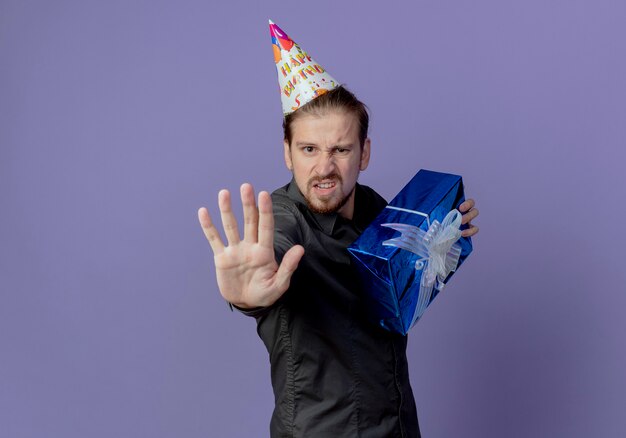 Free photo greedy handsome man in birthday cap stands sideways holding gift box and gesturing stop hand sign isolated on purple wall