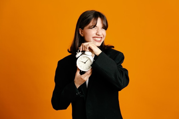 Greedy holding alarm clock young beautiful female wearing black jacket isolated on orange background