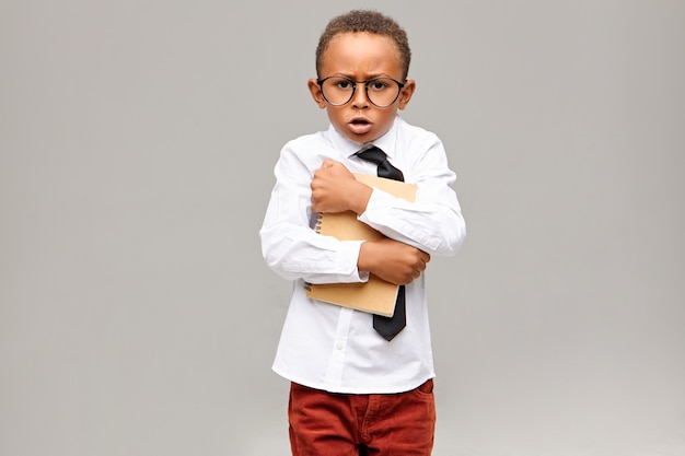 greedy nerdy dark skinned elementary pupil wearing school uniform and eyeglasses holding tight copybook and exclaiming. Childhood, education, learning and lifestyle concept