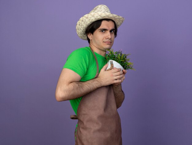 Greedy young male gardener in uniform wearing gardening hat holding flower in flowerpot