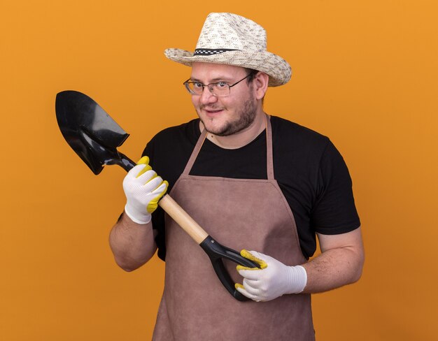 オレンジ色の壁に分離されたスペードを保持しているガーデニング帽子と手袋を身に着けている貪欲な若い男性の庭師