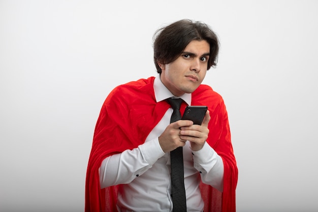 Greedy young superhero guy looking at camera wearing tie holding phone isolated on white