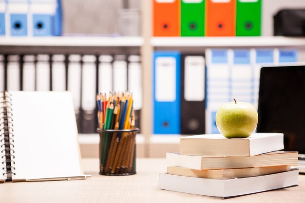 ノートブックの横にある本の山に青リンゴ、後ろにぼやけたホワイトボードが付いたテーブルに鉛筆。学校のコンセプト