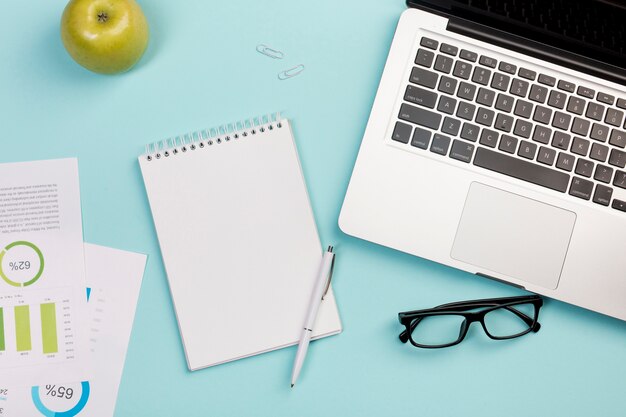 Green apple,spiral notepad,pen,eyeglasses and laptop on blue background