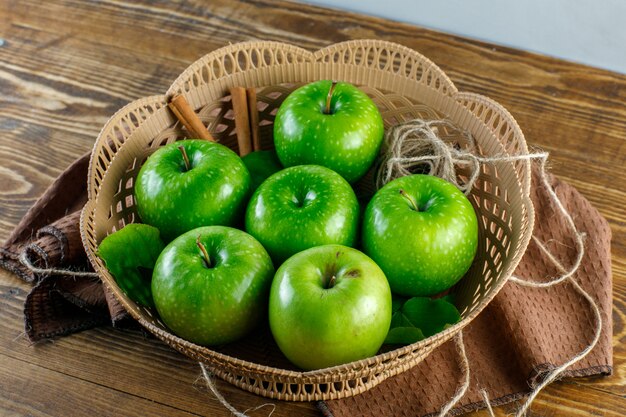 Free photo green apples in a basket with cinnamon sticks, rope, kitchen  towel, leaves high angle view on wooden table