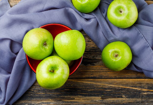 Green apples in a bowl on wooden and textile background, flat lay.