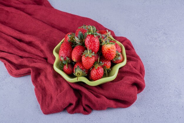 Green bowl of strawberries nested on a wrinkled pile of tablecloth on marble background. High quality photo