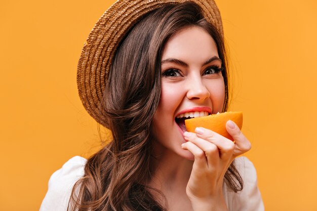 Free photo green-eyed brunette girl in straw hat bites juicy orange on isolated background.