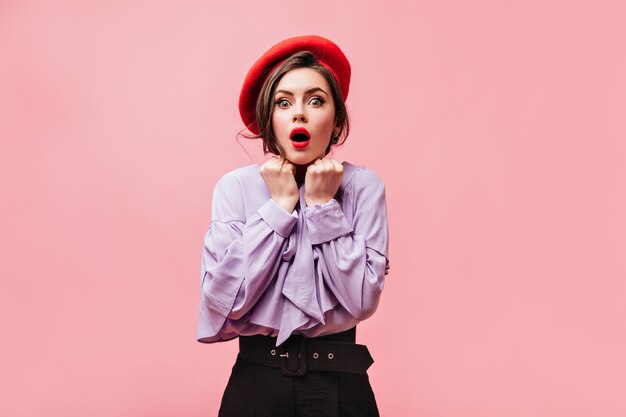 Green-eyed brunette woman in bright beret and blouse with fright looks into camera on pink background.