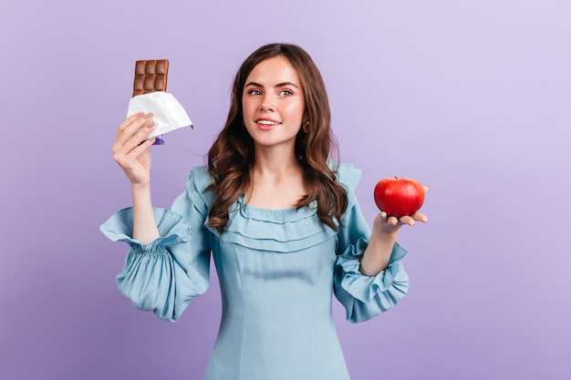 Green-eyed curly brunette girl posing with red apple and bar of milk chocolate on purple wall.