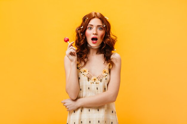 Green-eyed curly woman in checkered sundress holds pink lollipop and stares in surprise at camera on orange background.