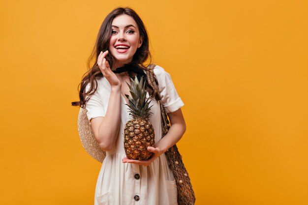 Green-eyed girl in white dress holding pineapple. Woman laughs and poses with shopping bag on orange background.