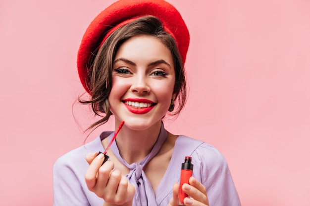 Free photo green-eyed girl with snow-white smile paints her lips with red lipstick. portrait of lady in stylish beret on pink background.