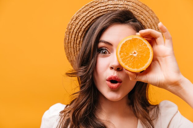 Green-eyed girl with wavy hair looks at camera in surprise and covers her eye with orange.
