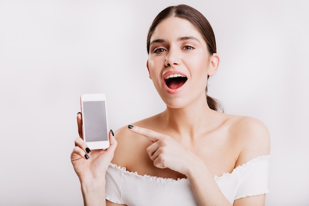 Green-eyed girl without makeup demonstrates phone on white wall.
