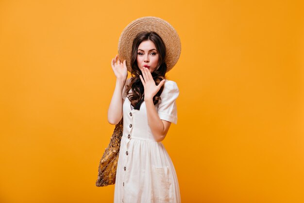 Green-eyed lady covers her mouth with her hand. Woman in straw hat and white sundress holds shopping bag on orange background.