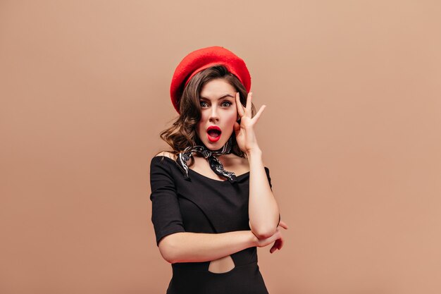 Green-eyed lady with red lips looks into camera in surprise and touches her face. Shot of brunette in stylish beret and elegant dress.