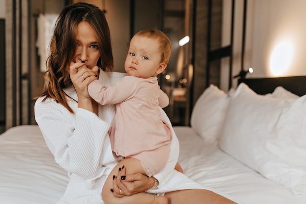 Green-eyed woman in bathrobe kisses hand of her child. Mom and daughter pose at home sitting on bed.