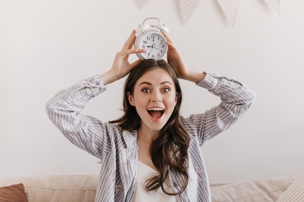 Green-eyed woman enthusiastically looking at front and holding alarm clock on her head