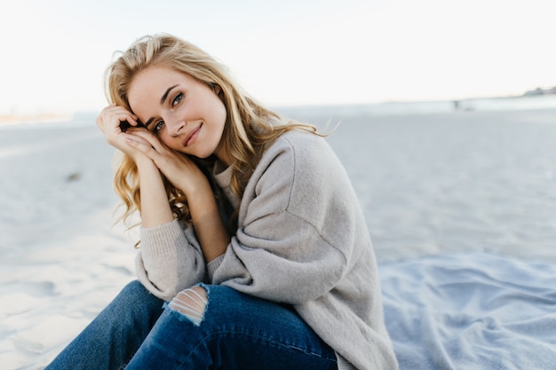 Green-eyed woman in jeans and sweater is sitting in sand, leaning in her knees in the sea.