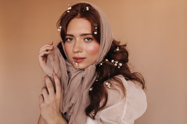 Green-eyed woman in light outfit coquettely looks at camera on beige background, playing with scarf.