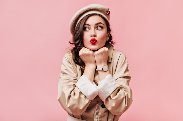 Green-eyed woman in trench coat and hat folded her lips into tube and posing on isolated background.