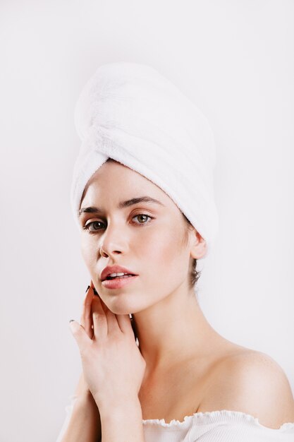 Green-eyed woman with perfect skin posing on white wall with towel on her head.