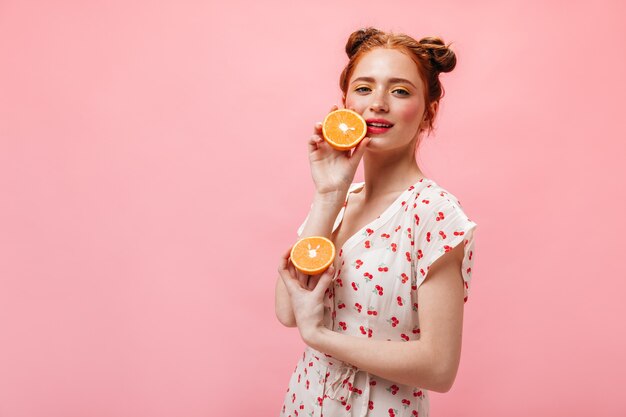 La donna dagli occhi verdi con i capelli rossi guarda con stupore la telecamera e tiene succose arance su sfondo rosa.